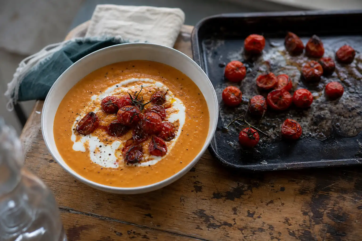Djup skål med rödorange linssoppa toppad med rostade, lätt brända tomater och yoghurt, bredvid en sotig plåt med rostade tomater på ett slitet träbord.