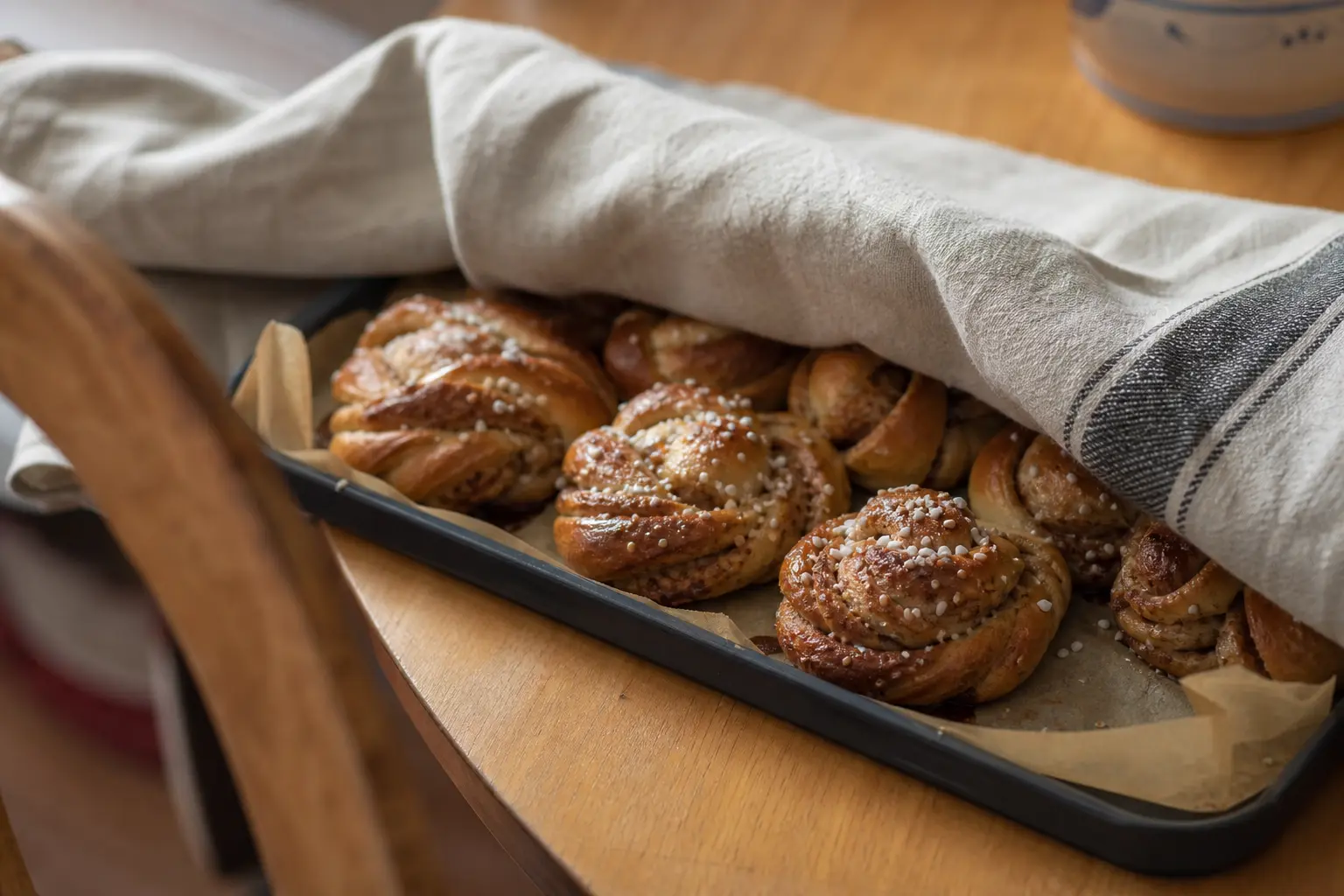 Nygräddade kanelbullar med pärlsocker på plåt, med en bakduk halvt dragen över.