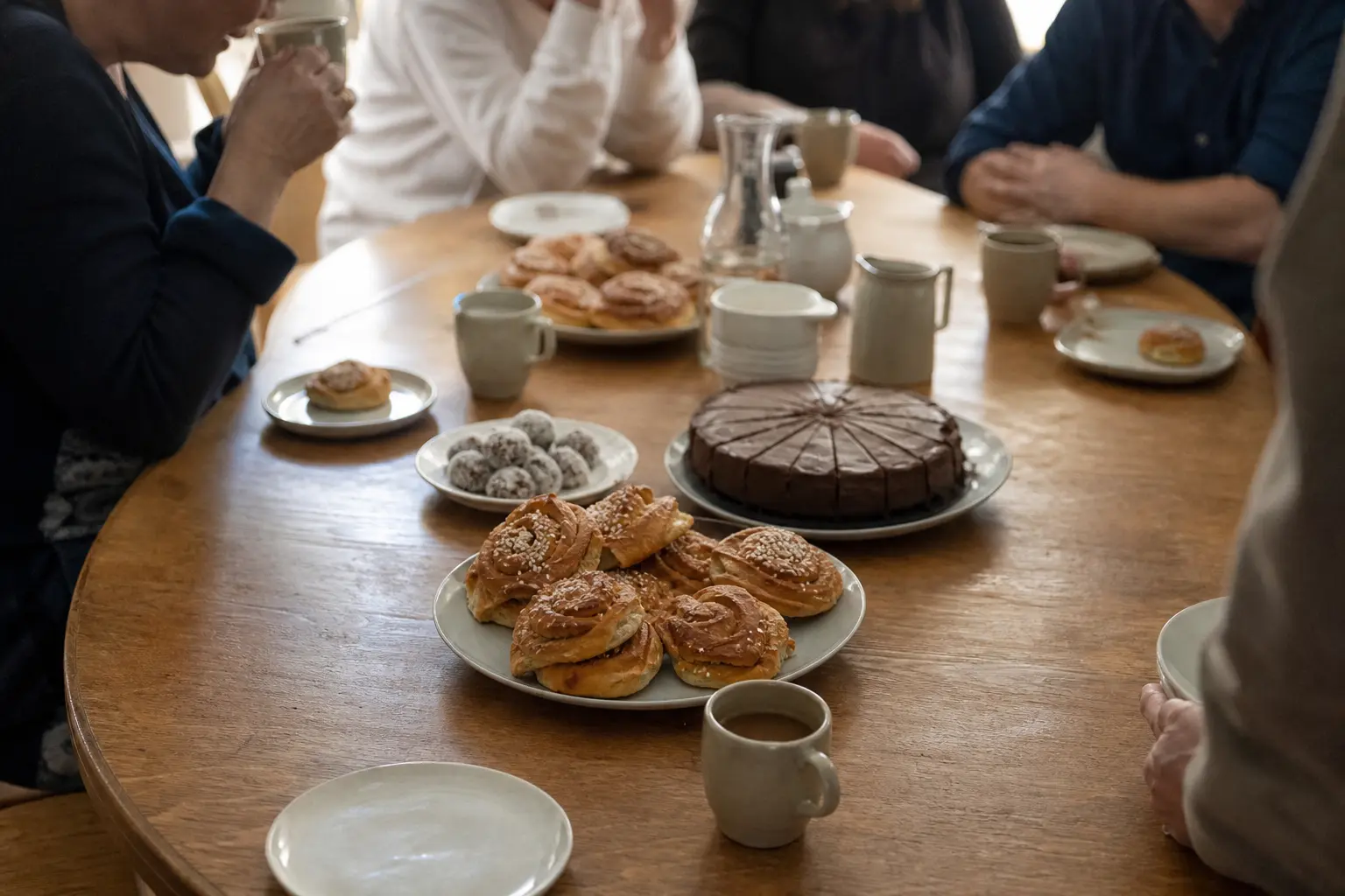 Runt träbord med kanelbullar, uppskuren chokladkaka, chokladbollar och kaffekoppar, omgivet av personer som fikar.
